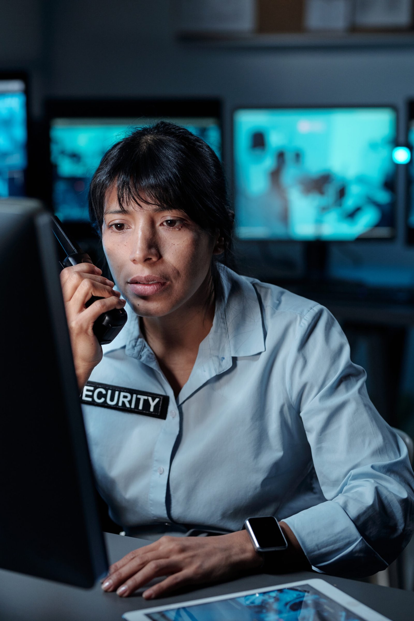 Young female patrol staff in uniform reporting situation on walkie-talkie and looking at computer screen while sitting by workplace